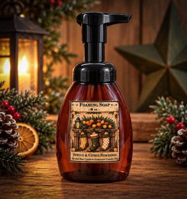 Spruce & Citrus Stockings 12 oz Foaming Hand Soap in an amber pump bottle with a vintage Christmas stocking label, displayed on a rustic wooden table with festive greenery, pinecones, and warm holiday lights.