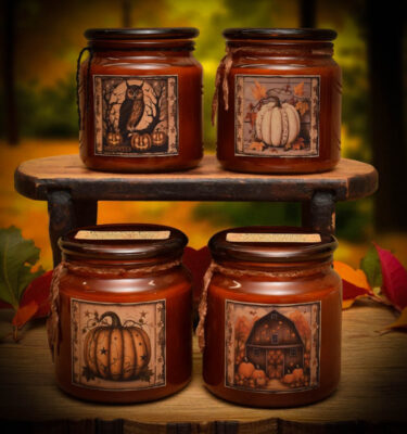 Four amber glass fall candles with folk art labels featuring an owl, white pumpkin, orange pumpkin, and rustic barn, displayed on a wooden shelf with autumn leaves.