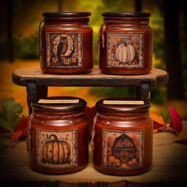 Four amber glass fall candles with folk art labels featuring an owl, white pumpkin, orange pumpkin, and rustic barn, displayed on a wooden shelf with autumn leaves.