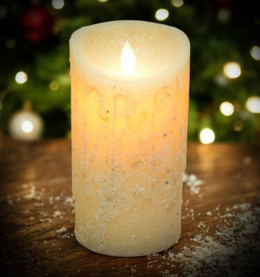 Drizzled snowy white moving flame pillar candle glowing with warm light, displayed on a rustic wood surface with Christmas lights softly blurred in the background.