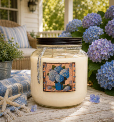 Heavenly Hydrangea 64 oz two-wick floral candle displayed on a coastal cottage porch surrounded by blooming blue hydrangeas.