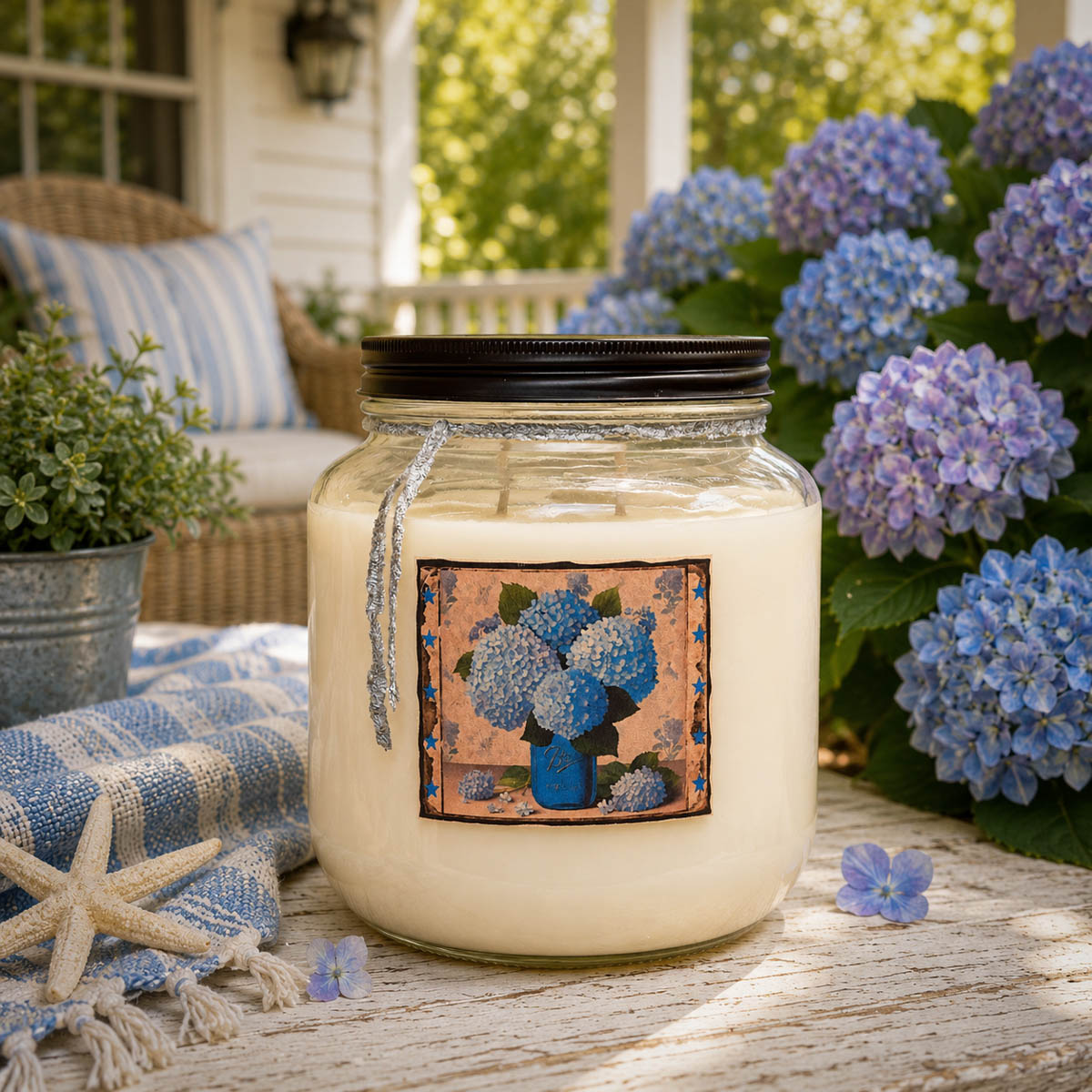 Heavenly Hydrangea 64 oz two-wick floral candle displayed on a coastal cottage porch surrounded by blooming blue hydrangeas.